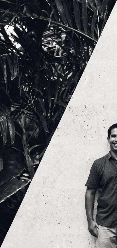 DC Chavez smiling, leaning against a pale stone wall beside dark palm fronds